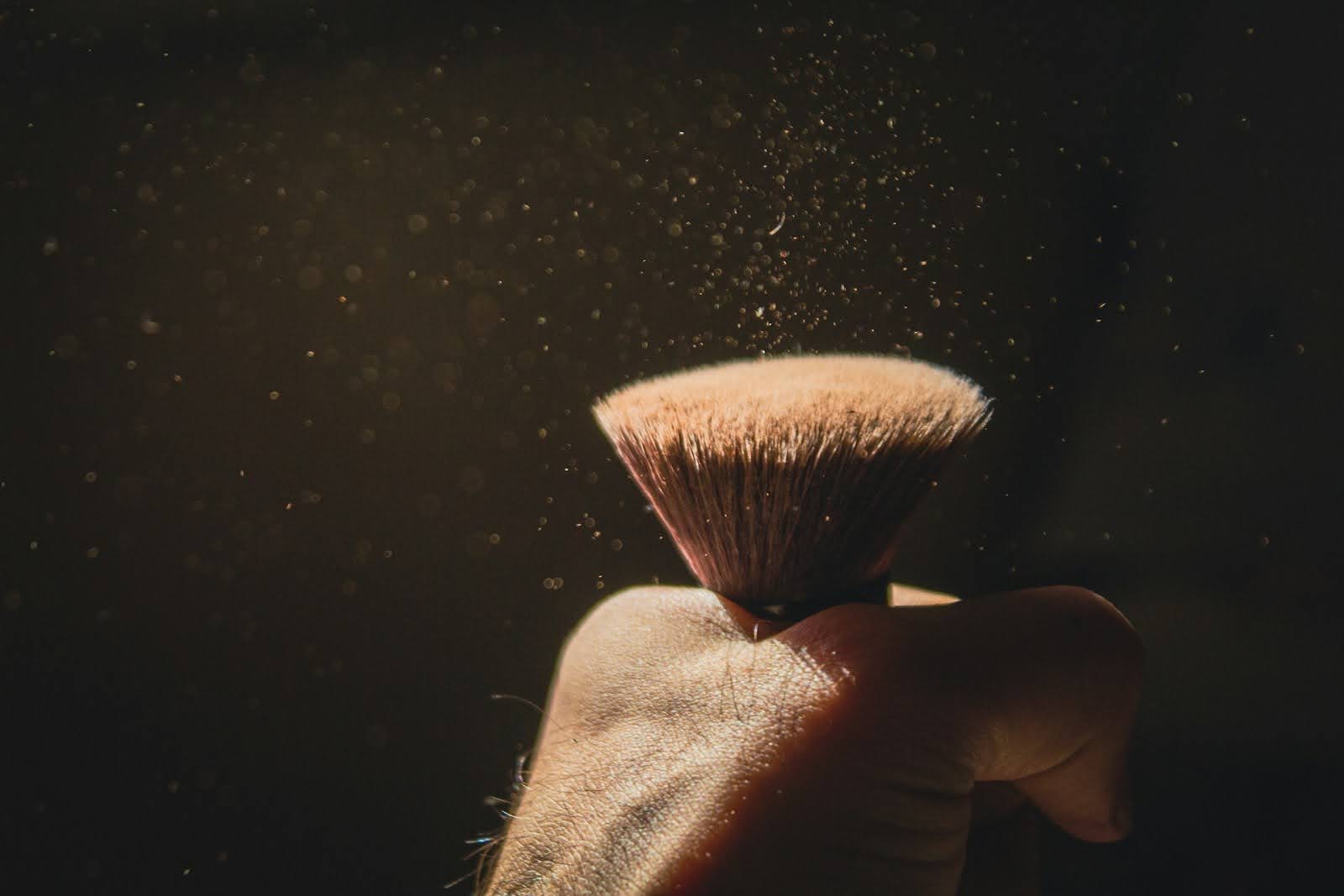 Hand holding a makeup brush as loose powder disperses into the air against a dark background.