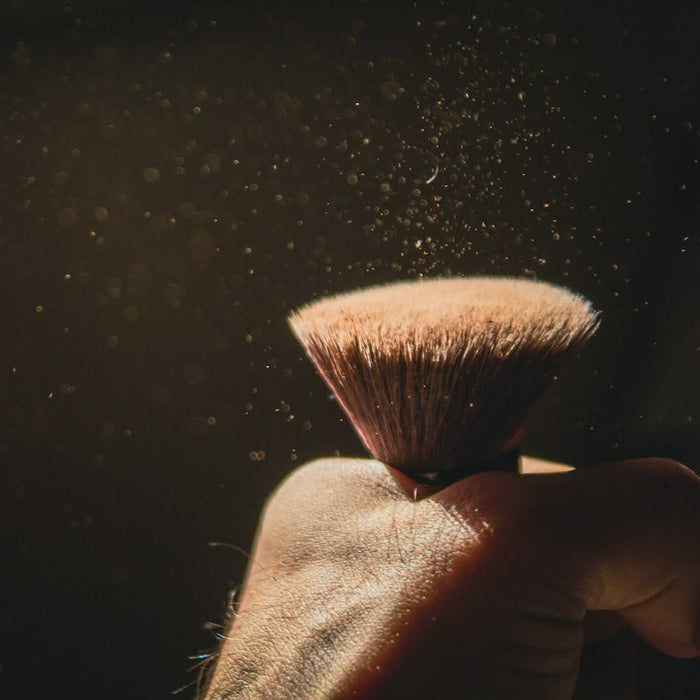 Hand holding a makeup brush as loose powder disperses into the air against a dark background.
