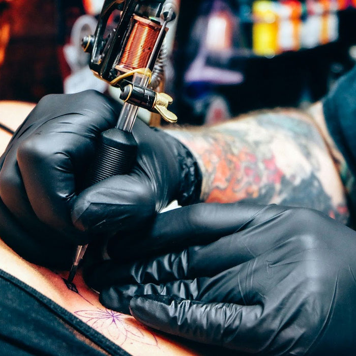 Tattoo artist wearing black gloves applying ink to skin with a tattoo machine in a studio setting