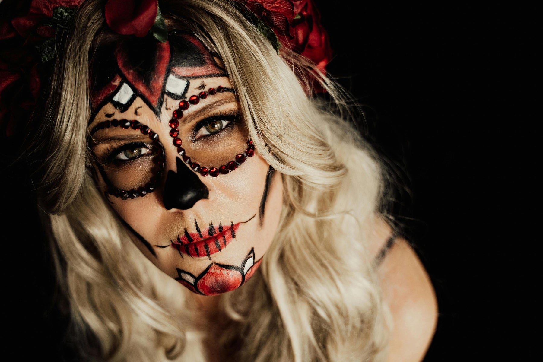 Woman with detailed sugar skull face paint, red gemstone accents, and a red floral headpiece against a dark background